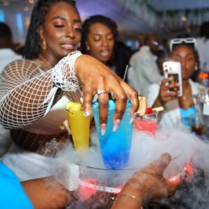 A lady reaching into the foreground to grab a blue cocktail at MUKE Life's All White Soiree